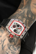 A close-up of a wristwatch with a silver-tone case, black strap, and a white dial accented with red trim and black subdials. The watch is shown worn on a tattooed arm against a black background.