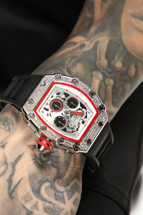 A close-up of a wristwatch with a silver-tone case, black strap, and a white dial accented with red trim and black subdials. The watch is shown worn on a tattooed arm against a black background.