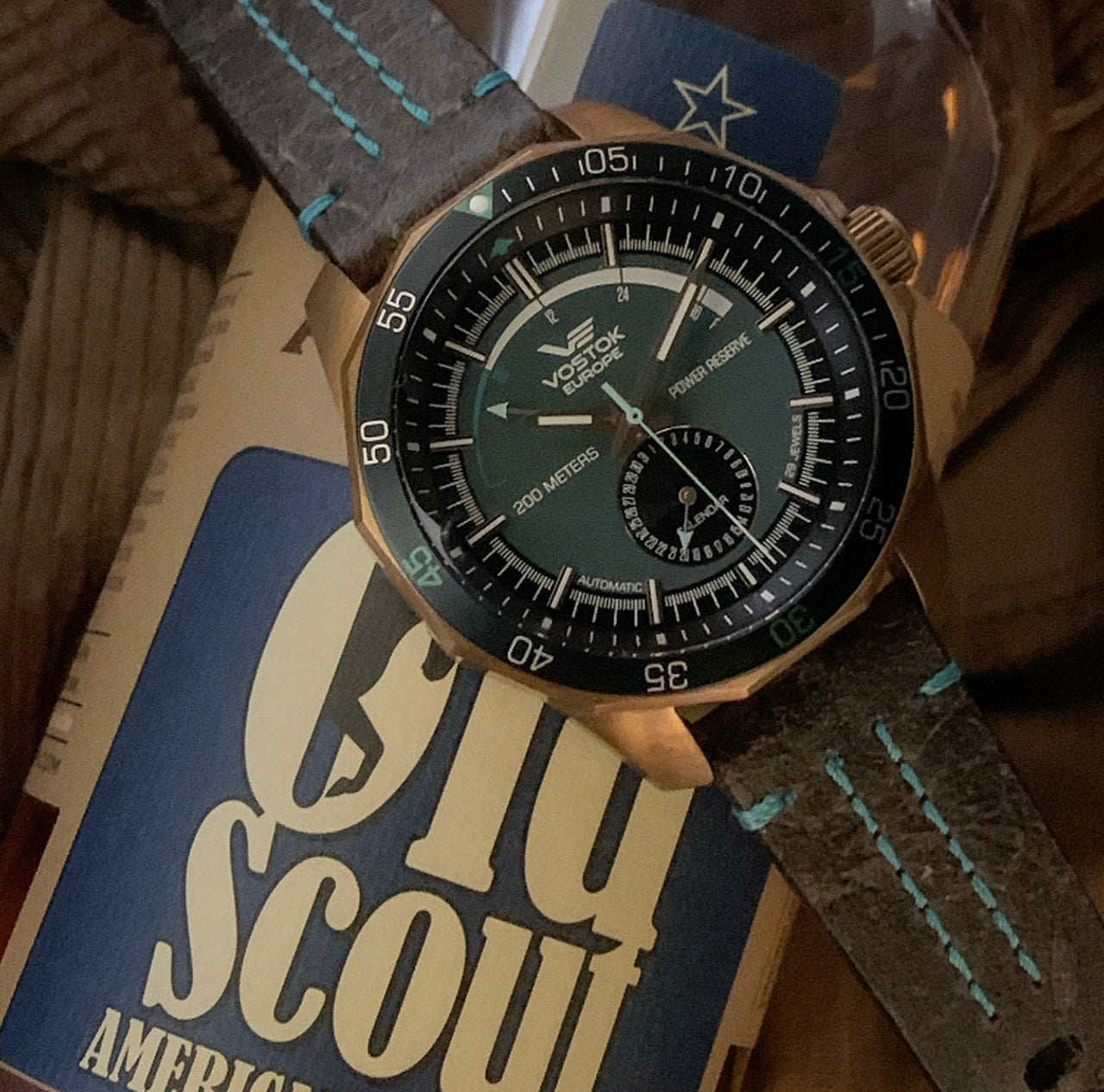 A close-up of a & Oak wristwatch with a dark green dial, bronze-toned case, and dark gray textured strap with turquoise stitching.
