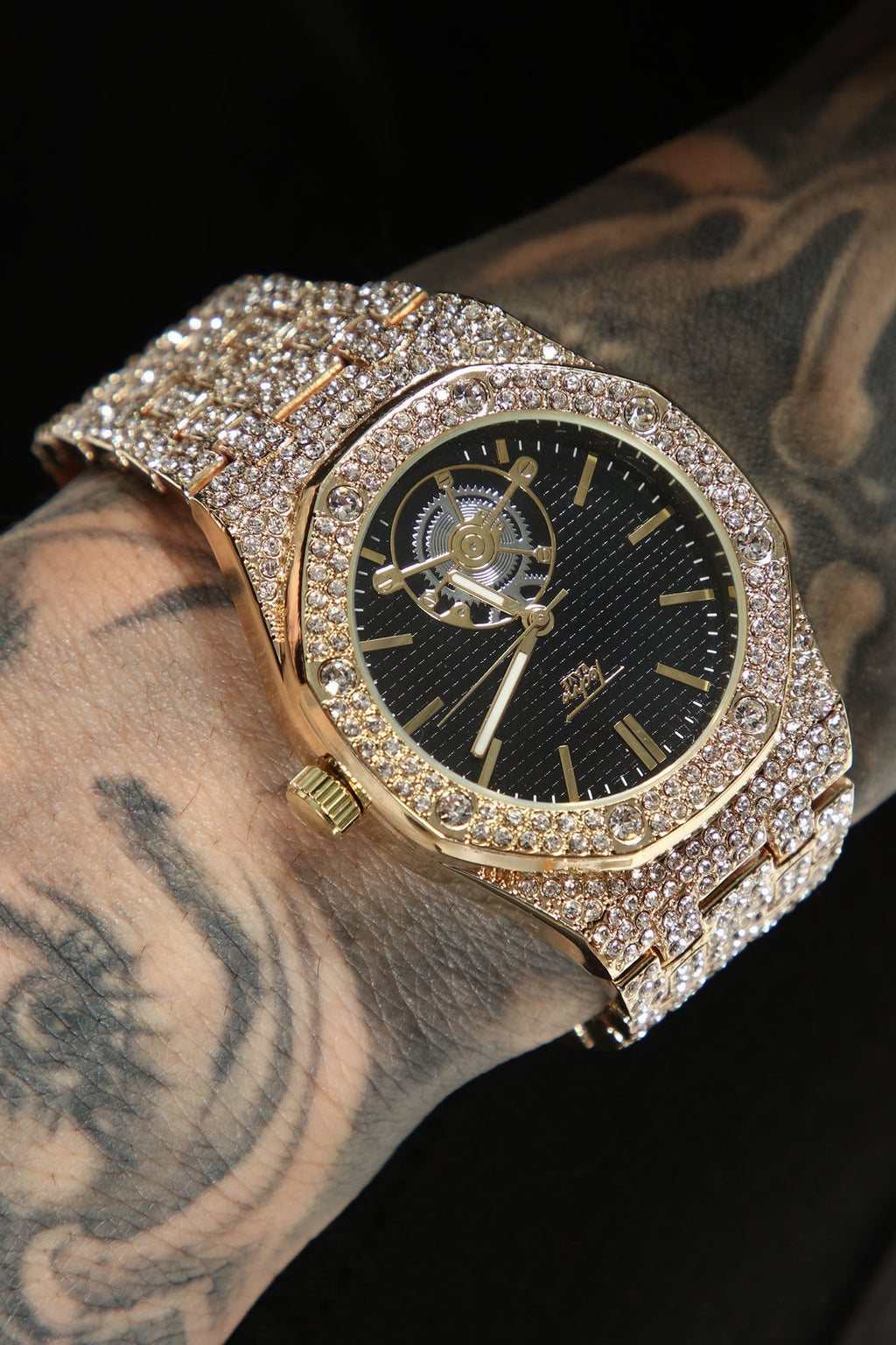 A gold-tone wristwatch heavily embellished with clear rhinestones, shown worn on a tattooed wrist against a black background.