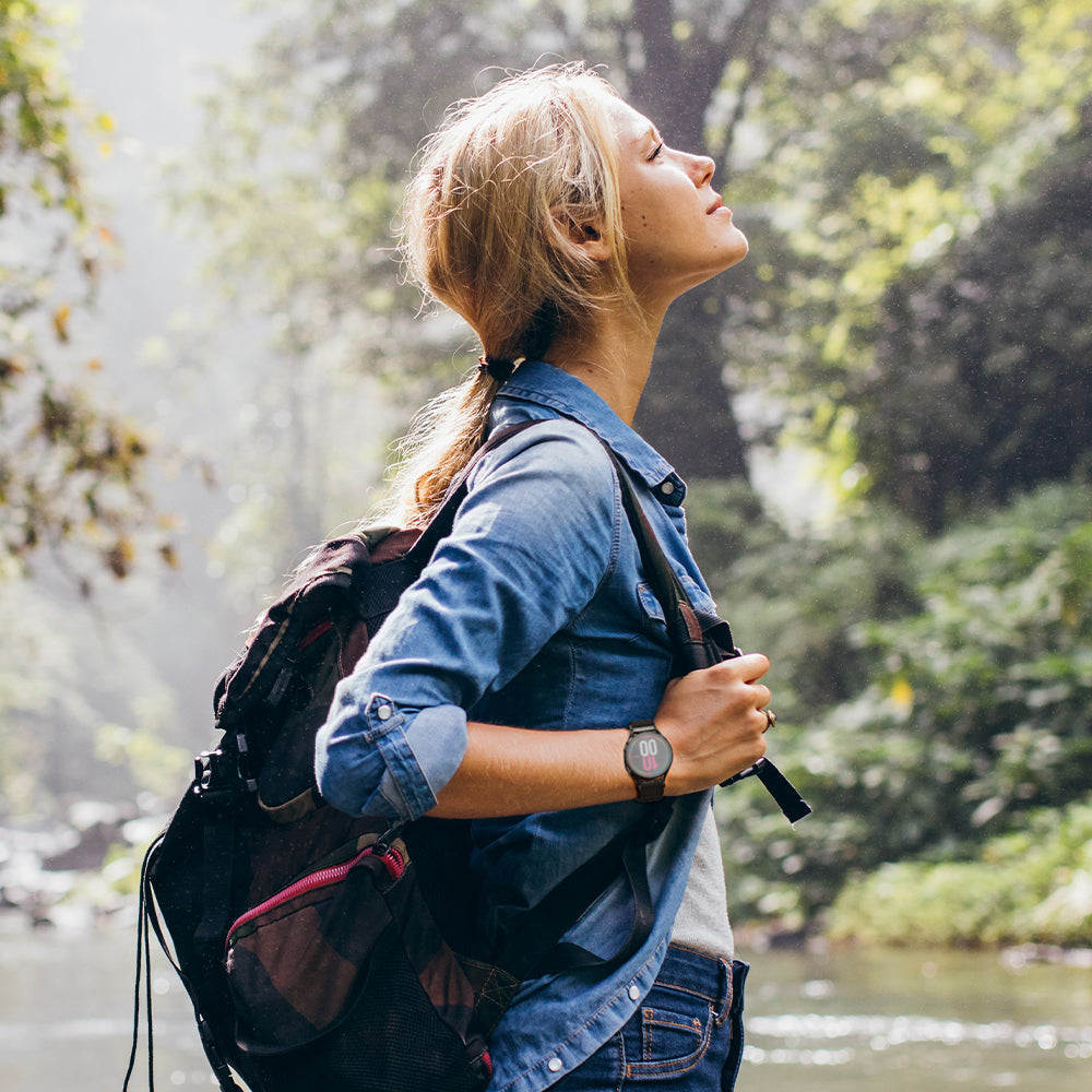 A woman in a blue denim shirt and jeans hiking with a black and red backpack, looking up at trees near a stream.